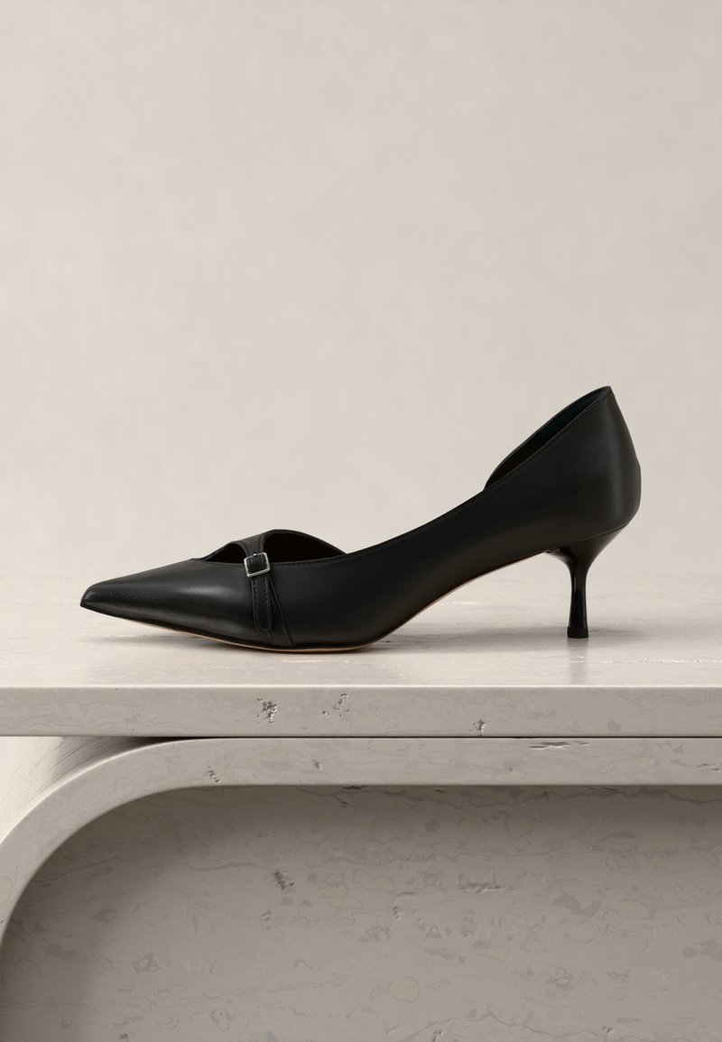 Μαύρο δερμάτινο παπούτσι με μυτερή μύτη και χαμηλό τακούνι, με μικρό λουράκι με πόρπη, τοποθετημένο πάνω σε ανοιχτό γκρίζο πέτρινο υπόστρωμα πάνω σε ουδέτερο φόντο.