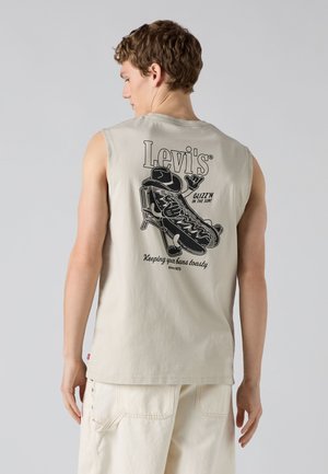 Homme portant un t-shirt beige sans manches Levi's avec un graphisme noir d'un personnage hotdog en train de se détendre et le texte "GLIZZ'N IN THE SUN!" dans le dos.
