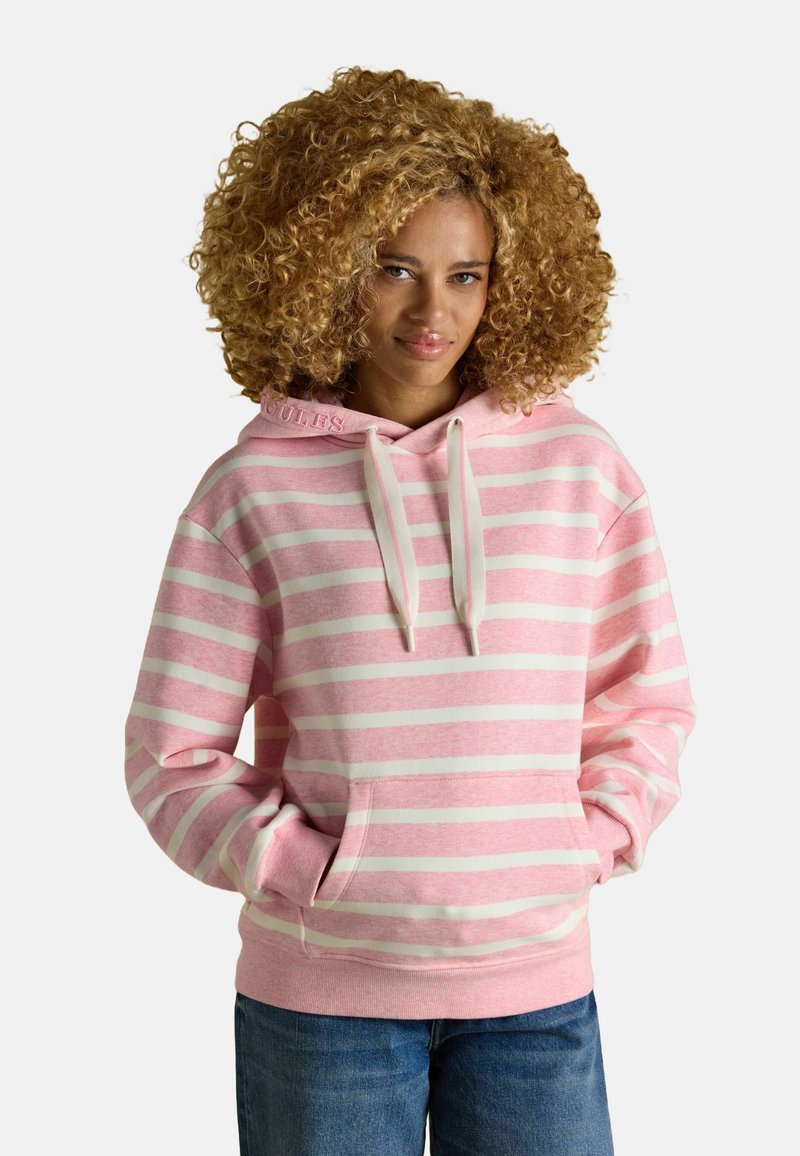 Žena s kučeravými vlasmi, oblečená v ružovo-bielom pruhovanom mikinovom svetri a modrých džínsoch, s rukami v vreckách mikiny, na jednoduchom pozadí.