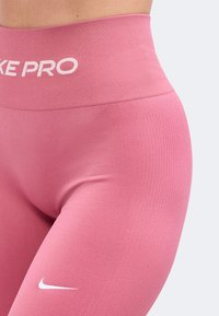 Leggings Nike Pro roses taille haute avec logo blanc sur la taille et petit swoosh Nike sur la cuisse, portés sur le bas du torse et les jambes d'une personne.