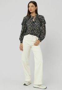 Femme portant un blouse à volants florale noire avec des manches bouffantes, un pantalon large taille haute blanc et des baskets blanches, se tenant devant un fond gris.