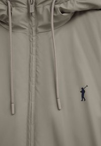 Beige jakke med forreste lynlås, snoretræk og lille mørkeblåt logo af en person, der holder en polo mallet på højre side.