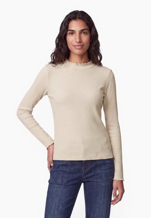 Donna con capelli scuri e ondulati che indossa una maglia beige a costine a maniche lunghe e jeans blu scuro, in piedi con una mano sui fianchi.