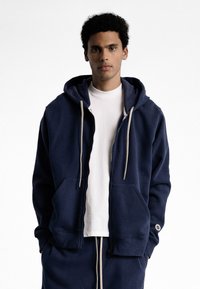 Hoodie zippé bleu marine en tissu doux avec une grande capuche, des poches avant et des cordons de serrage crème. Comprend des poignets et un ourlet côtelés.