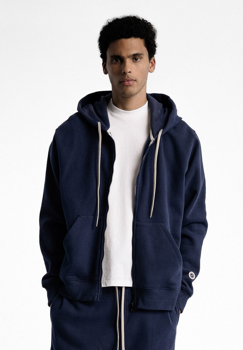 Hoodie zippé bleu marine en tissu doux avec une grande capuche, des poches avant et des cordons de serrage crème. Comprend des poignets et un ourlet côtelés.