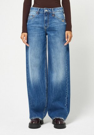 Personne debout portant un jean bleu à jambes larges, un haut marron foncé à manches longues et des chaussures marron foncé à semelle épaisse, sur un fond blanc.