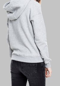 Persona vistiendo una sudadera gris claro y jeans negros, vista desde atrás contra un fondo gris liso.