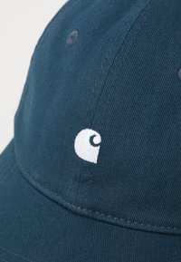 Mørkeblå caps laget av teksturert stoff, med en hvit brodert logo på forsiden. Capsen har en buet skygge og sydd detaljer.