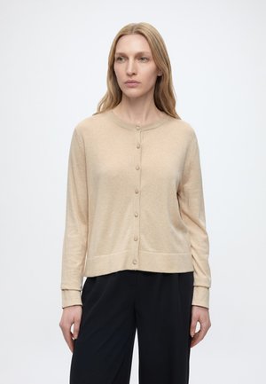 Femme aux longs cheveux blonds portant un cardigan beige boutonné et un pantalon noir, debout devant un fond clair uni.