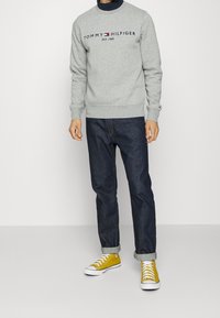 Sudadera gris con cuello redondo y logo "TOMMY HILFIGER". Vaqueros azul oscuro con puños doblados y zapatillas amarillas con suelas blancas.