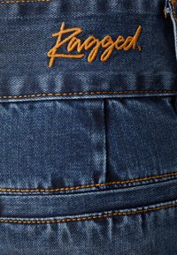 Dżinsowy materiał w ciemnoniebieskim odcieniu; z pomarańczowymi szwami i wyszywanym logo "Ragged"; tekstura klasyczna i lekko zużyta.