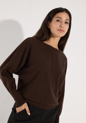 Vrouw met lang donker haar, die een losse donkerbruine trui en zwarte broek draagt en poseert met één hand in de zak tegen een effen achtergrond.