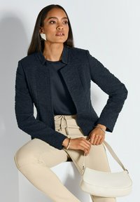 Marineblauwe gestructureerde jas met een kraag, gedragen met een donkere top en lichtbeige broek. Houdt een kleine crèmekleurige handtas vast, met een eenvoudig ontwerp.