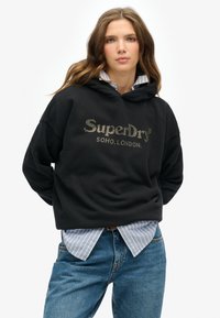 Felpa nera con testo "SuperDry" in dettaglio metallico. Indossata sopra una camicia a righe azzurre e bianche. Vestibilità casual, tessuto morbido.