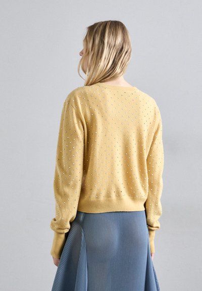 Κίτρινο πλεκτό πουλόβερ με υφή, χαρακτηριστικό για τις ανυψωμένες κουκίδες του. Φορεμένο πάνω από μια πλισέ μπλε φούστα, επιδεικνύοντας ένα τετράγωνο σχήμα και ριγωτούς μανίκες.