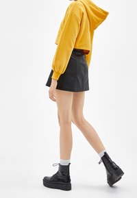 Persona vistiendo un hoodie cortado de color amarillo mostaza, una mini falda negra, calcetas blancas y botas de plataforma negras con cordones, caminando hacia la derecha.