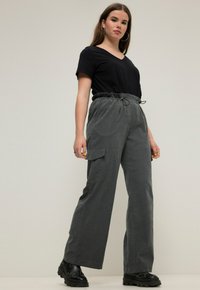 Harmaat cargo-housut, joissa on rento istuvuus, joustava vyötärönauha ja sivutaskut, yhdistettynä mustaan V-kaula-aukkopaitaan ja massiivisiin mustiin saappaisiin.