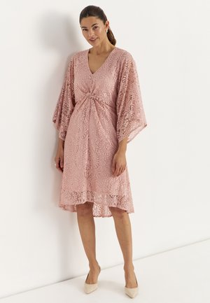 Kobieta w różowej, spranej koronkowej sukience do kolan z szerokimi rękawami i wiązanym detalem w talii, stojąca przy białej ścianie.