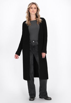 Frau, die einen langen schwarzen Cardigan, ein dunkelgraues Shirt, ausgebleichte schwarze Jeans und schwarze Stiefel trägt und vor einem weißen Hintergrund steht.