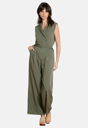 Kobieta o ciemnych włosach, ubrana w oliwkowo-zielony kombinezon bez rękawów z paskiem oraz beżowe sandały na koturnie z odkrytymi palcami, stojąca z jedną ręką w kieszeni.