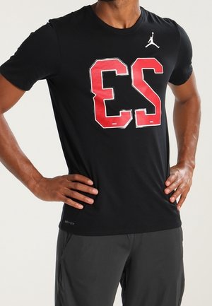 Homme en t-shirt sportif noir avec un grand numéro rouge 23 et un logo Jumpman blanc, les mains sur les hanches, portant un short sombre.
