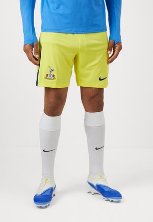 Gule sports shorts med en blå side stripe, der har et broderet logo. Parret med hvide knæstrømper og blå fodboldsko.