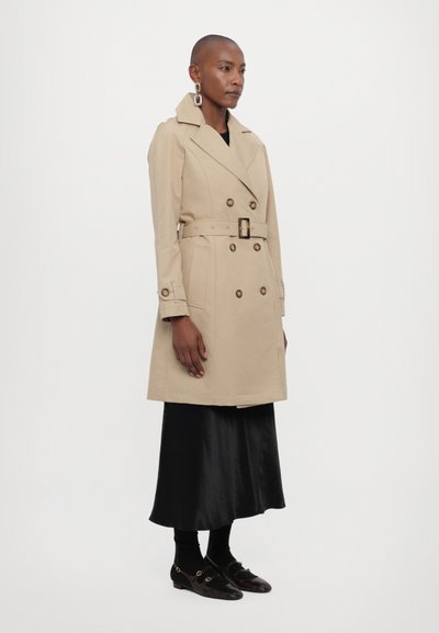 Beige trench coat with double-breasted buttons, a belt at the waist, and shoulder epaulettes, worn over a black satin skirt and stockings.