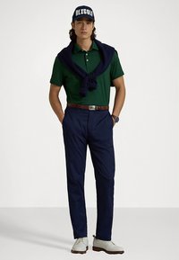 Grønn polo skjorte, marine blå bukser, marine blå genser drapert over skuldrene, hvitt belte, klokke og lyse sko. Blå caps med teksten "RLXGOLF".