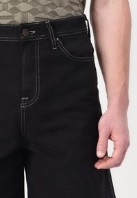 Pantalon en denim noir avec des coutures blanches, présentant des poches avant et une fermeture à bouton. Le tissu est robuste avec une coupe droite.