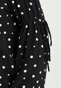 Black fabric blouse with white polka dots. Features gathered sleeves with drawstring accents for adjustable fit. Soft, textured material.