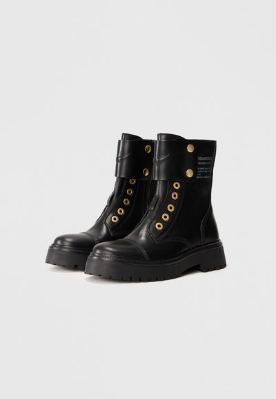 Black leather ankle boots with a chunky sole, featuring gold eyelets, a side zipper, and a snap closure at the cuff. Text on the back.