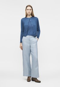 Žena v modrém zapnutém svetříku přes ladící top, světle modrých širokých kalhotách a hnědých kotníkových botách, stojící na bílém pozadí.