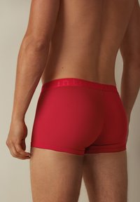 Röda boxertrosor i slät tyg, med en bred elastisk midjeband som har varumärkesnamnet, och en åtsittande design som formar sig efter kroppen.