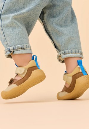 Kind trägt beige-braune Schuhe mit weicher Sohle und blauen Zuglaschen, geht vorsichtig mit hochgekrempelten hellblauen Jeans vor einem beigen Hintergrund.