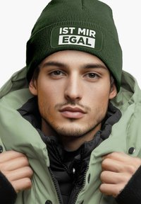 Green knit beanie with a white patch reading "IST MIR EGAL," worn with a light green puffer jacket and a black zipper jacket underneath.