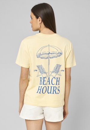 Femme aux longs cheveux bruns portant un t-shirt jaune avec l'inscription « Beach Hours » et un motif d'ombrelle et de chaises longues, associé à un short blanc.