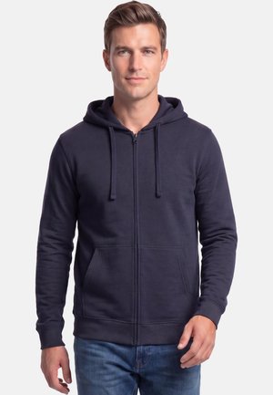 Man draagt een marineblauwe zip-up hoodie met trekkoorden en een voorzak, gecombineerd met blauwe jeans, staand tegen een witte achtergrond.