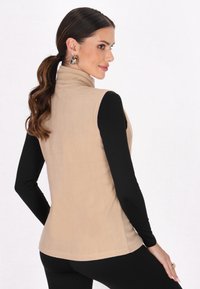 Beige Fleece-Weste mit hohem Kragen, glatter Textur und ärmellosem Design, kombiniert mit einem schwarzen Langarmshirt und eng anliegenden Hosen.