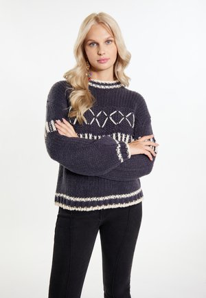 Pulover tricotat de culoare gri închis, cu o textură groasă, prezentând modele geometrice albe și manșete și tiv cu dantelă. Purtați cu pantaloni negri mulati.