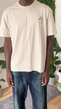 Beige Baumwoll-T-Shirt mit rundem Halsausschnitt, versehen mit einem kleinen grünen Palmenmotiv auf der linken Brust. Kombiniert mit lockeren dunkelblauen Jeans.