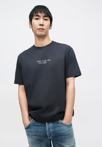 T-shirt bleu marine foncé à manches courtes avec col rond ; affiche le texte "GOOD TIMES AND TAN LINES" en blanc, assorti à un jean bleu clair déchiré.