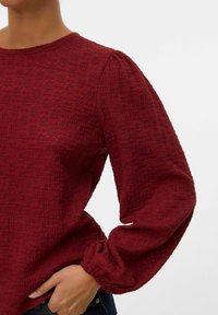 Bordeauxfärgad texturerad blus med långa puffsleeves, som har rund halsringning och rynkade manschetter. Tyget har ett subtilt vävt mönster.
