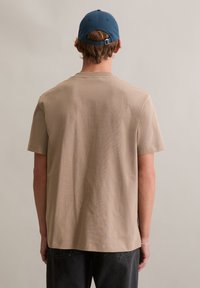 Beżowa bawełniana koszulka z krótkim rękawem, o okrągłym dekolcie i luźnym kroju, noszona z niebieską czapką z daszkiem. Prosty i nieozdobiony wzór.