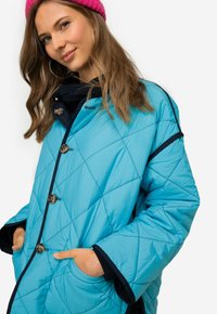 Vadderad blå pufferjacka med stora bruna knappar, svart kantning och sidofickor; har en något oversized design och mjuk textur.