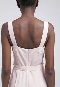 Vaaleanpunainen hihaton mekko, jossa on vartalonmyötäinen yläosa, teksturoitu kangas ja takana vetoketju. Mekossa on vyötäröä korostava vyö, joka on samanvärinen.