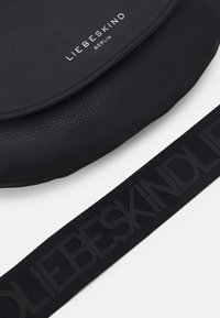 Borsa in pelle nera con una texture liscia, dotata di chiusura a pattina, logo del marchio in rilievo e tracolla in tessuto nero con stampa del logo.