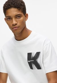 Jauns vīrietis ar īsiem izlocītiem matiem, tērpies baltā T-kreklā ar melnu izkropļotu "K" grafiku un tekstu "Karl Lagerfeld Jeans".