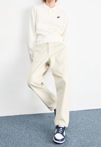 Ljusbeige långärmad pikétröja med svart Nike-logotyp, kombinerad med ljusbeige byxor och marinblå och vita sneakers. Slät tyg, avslappnad passform.