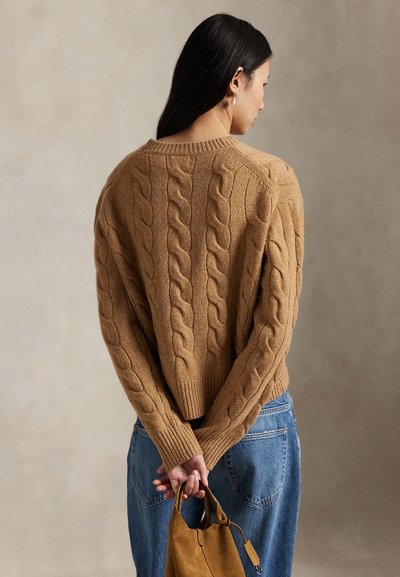 Žena oblečená do béžového kabelového svetr a modrých džín, držící hnědou semišovou kabelku za zády, obrácená zády k neutrálnímu pozadí.