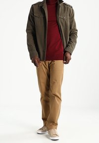 Olivengroene jas met knoopdetails, bordeaux trui, beige broek en lichte sneakers. Gestructureerde stoffen met een casual design.